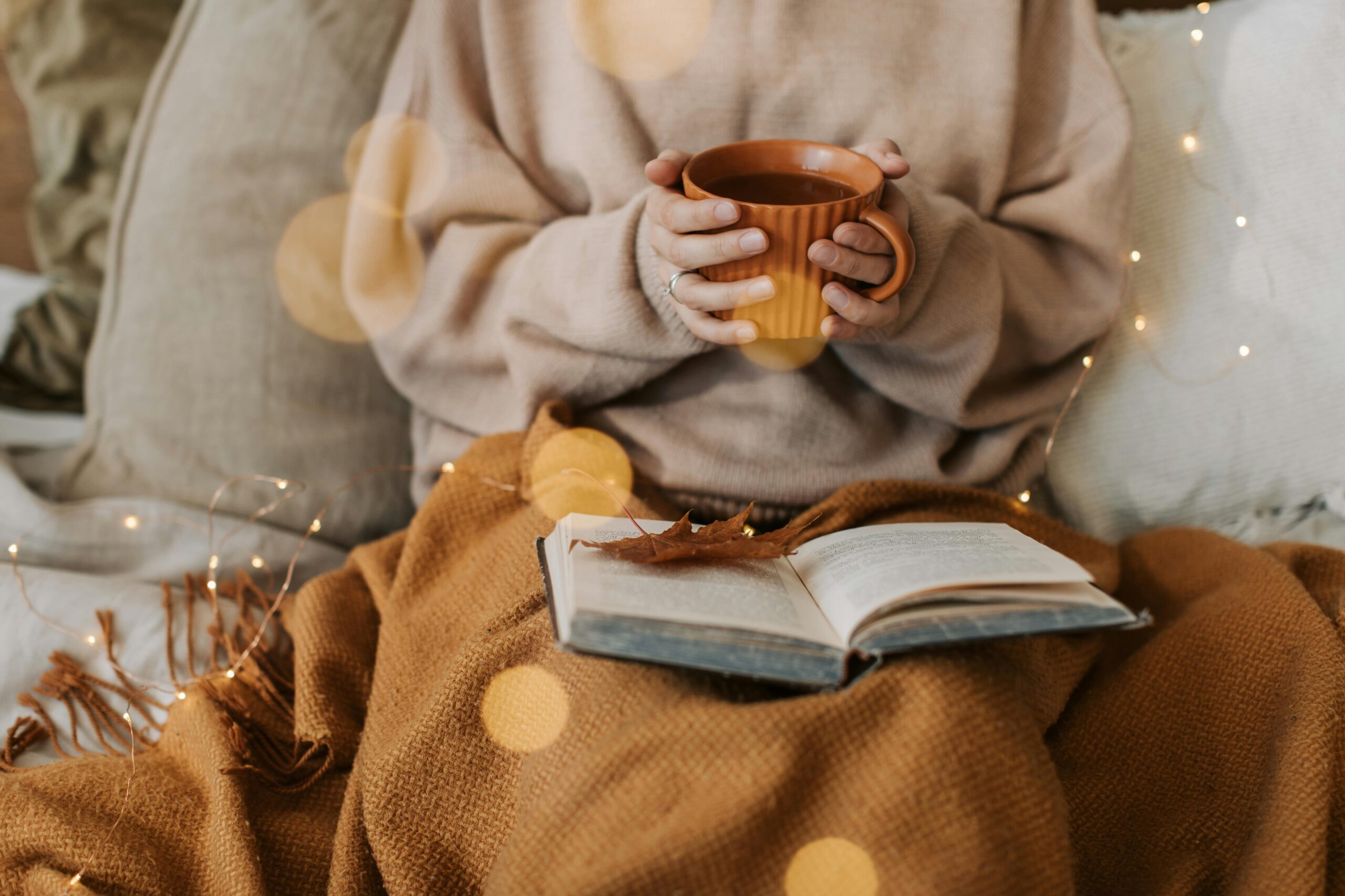 Warm autumn light, hands holding a cup of tea over an open book and cozy blanket — a gentle, quiet moment of presence and emotional calm.