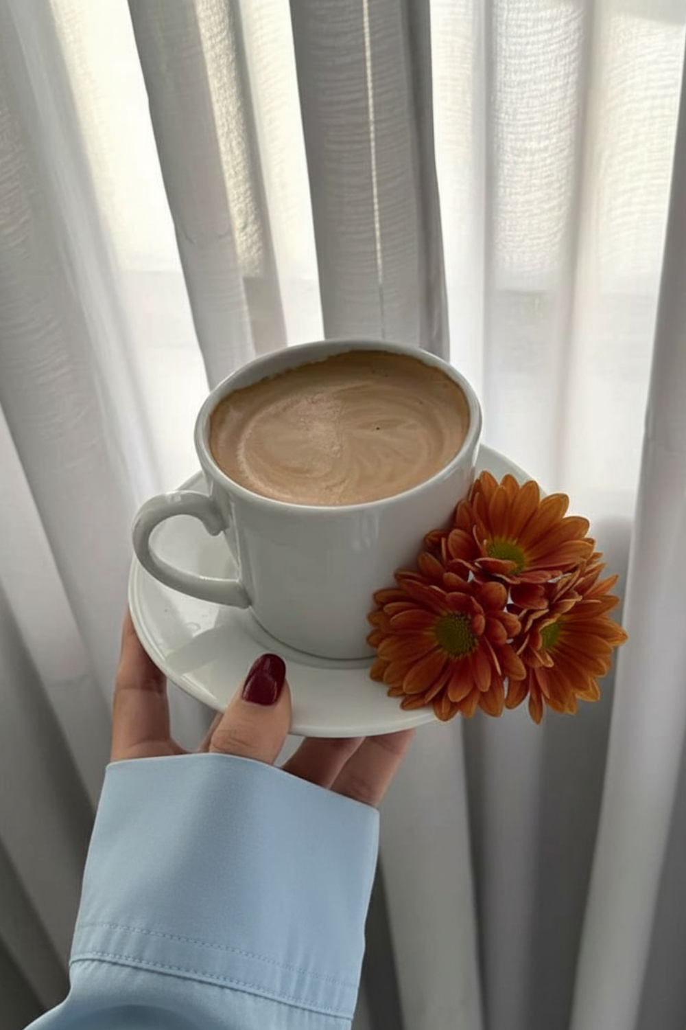 A hand holding a white coffee cup with a latte, framed by soft morning light through sheer curtains. A small orange flower rests on the saucer, creating a calm, cozy morning aesthetic.
