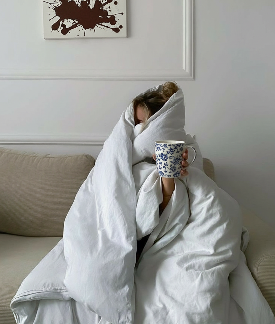 A woman wrapped in a white duvet, holding a coffee mug in a quiet morning moment — a soft, tired scene that reflects emotional exhaustion and slow returning to herself.