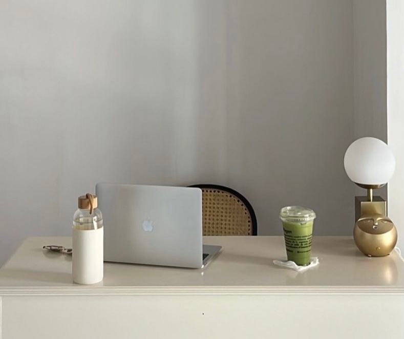A quiet, minimal work desk with a laptop, a glass water bottle, a matcha drink, and a small lamp on a clean surface. Soft light and empty space create a calm, tired-but-steady 9–5 atmosphere, reflecting the feeling of needing a quiet moment during the workday.