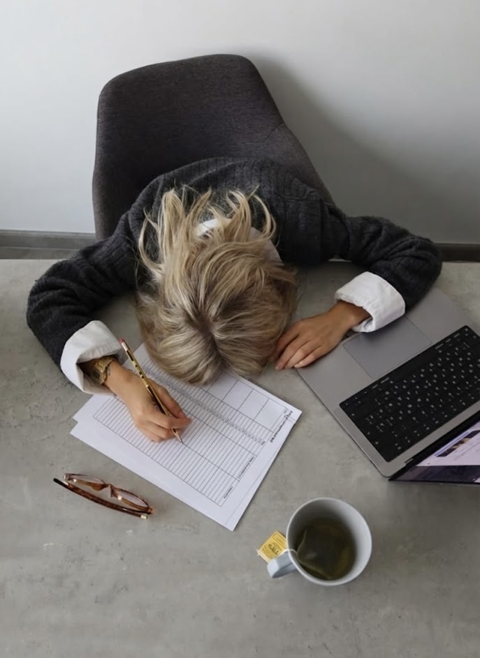 Exhausted woman with her head down on a desk, holding a pencil over unfinished paperwork next to a laptop and cup of tea, capturing the quiet burnout and overapologising energy of modern office life.