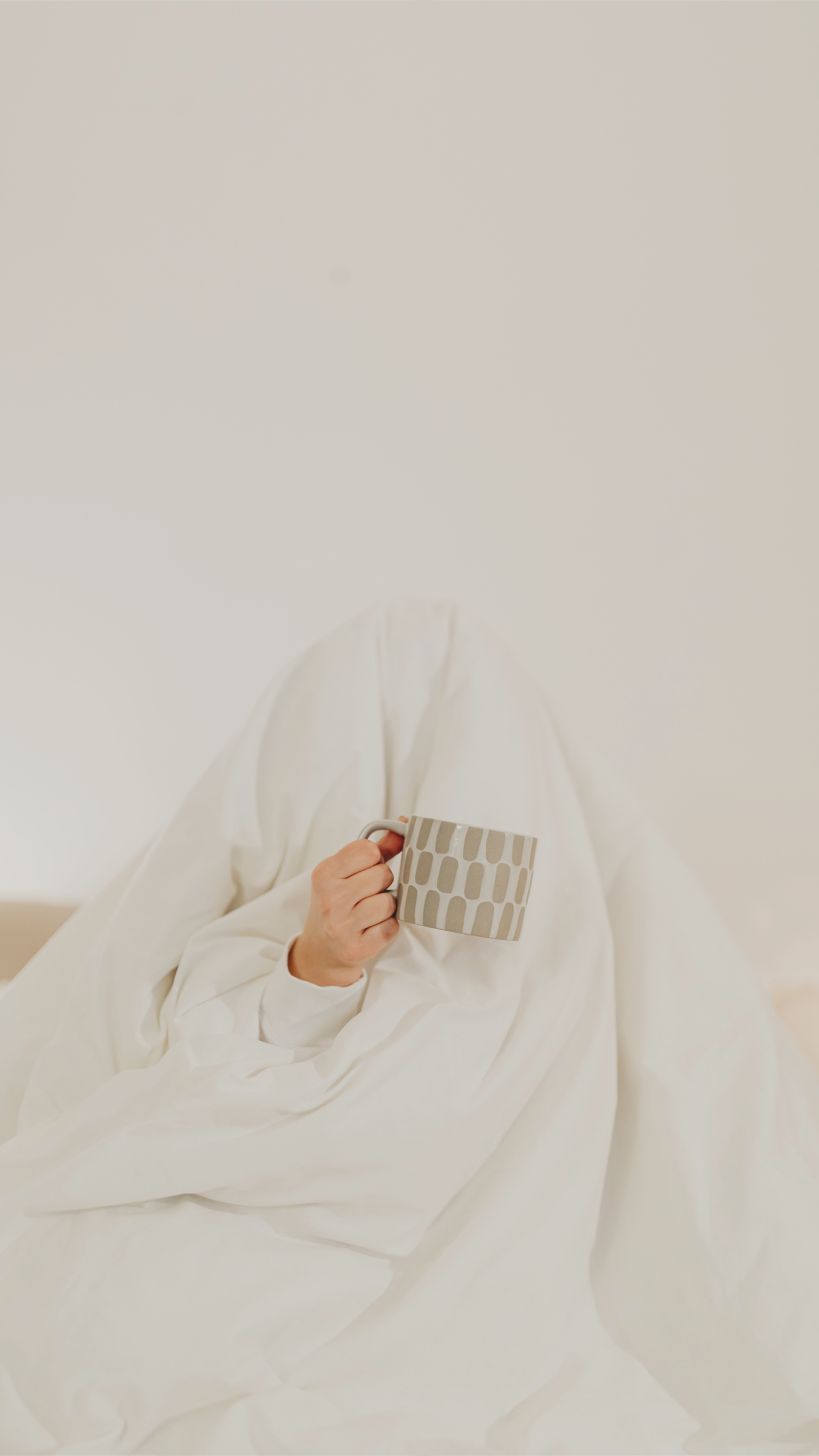 Person wrapped in a white blanket, holding a mug, sitting quietly in a minimal room. A calm, non-Instagram evening moment focused on rest, silence, and slow living without posting.