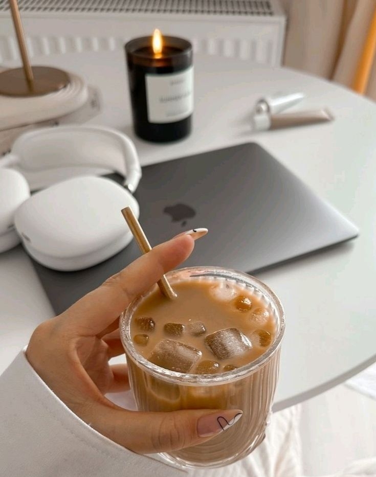 Woman holding an iced coffee in a cozy neutral home office setup with laptop, headphones and candle, representing calm productivity and focus planning.